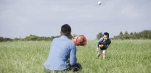 夕暮れ時の公園や庭で、父親と子どもがキャッチボールをする様子。 柔らかな夕日の光が差し込み、両者の表情が優しく照らされている。 父親は仕事帰りを思わせるビジネスカジュアル（Yシャツの袖をまくり、ネクタイを緩めた状態）。 子どもは真剣な表情でボールをキャッチしようとしている瞬間。